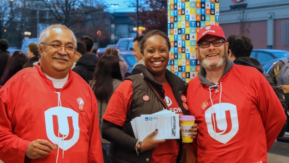 « Trois membres d’Unifor brandissent un drapeau Unifor en souriant à l’extérieur d’un arrêt de bus très fréquenté. »