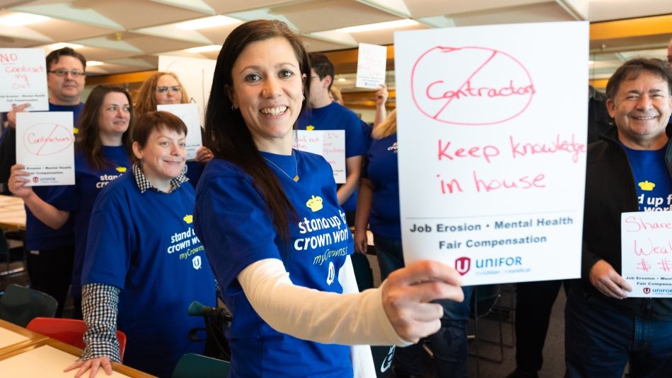 Une femme souriante en tee-shirt bleu tient une petite pancarte sur laquelle on peut lire "Pas de sous-traitance ; garder le savoir à l'intérieur". D'autres collègues portant des chemises bleues à l'arrière-plan. 